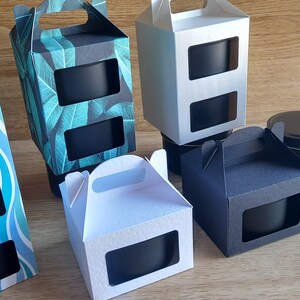 May include: Assortment of gift boxes with window cutouts, featuring different patterns and colors. The boxes are designed to hold candles or other small gifts. Some boxes have handles.