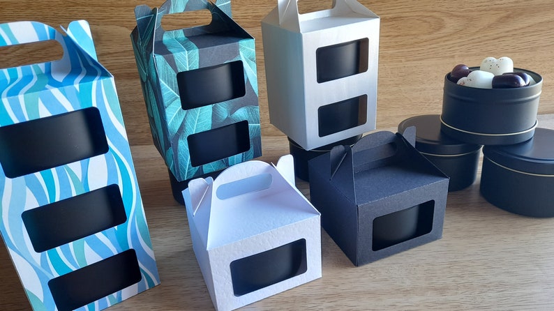 May include: Assortment of gift boxes with window cutouts, featuring different patterns and colors. The boxes are designed to hold candles or other small gifts. Some boxes have handles.