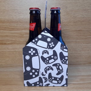 K&ouml;nnte beinhalten: Ein Sechs-Pack-Biertr&auml;ger mit wei&szlig;em Hintergrund und einem Muster aus schwarzen Videospiel-Controllern.