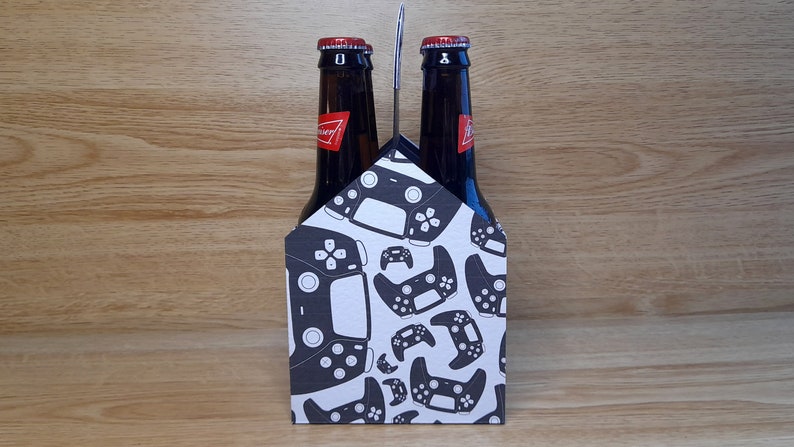 K&ouml;nnte beinhalten: Ein Sechs-Pack-Biertr&auml;ger mit wei&szlig;em Hintergrund und einem Muster aus schwarzen Videospiel-Controllern.