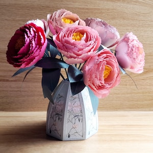 Può includere: Un bouquet di fiori di carta rosa e rossi in un vaso di ceramica bianca con un motivo floreale. Il vaso ha una forma geometrica con una base larga e un collo stretto.