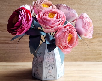 Könnte beinhalten: Ein Strauß aus rosa und roten Papierblumen in einer weißen Keramikvase mit Blumenmuster. Die Vase hat eine geometrische Form mit einem breiten Boden und einem schmalen Hals.
