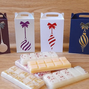 Puede incluir: Cuatro cajas decorativas con un diseño de adorno navideño en morado, rojo, blanco y dorado. Las cajas son de cartón y tienen un diseño recortado. Las cajas están sobre una superficie de madera con varias ceras derretidas delante de ellas.