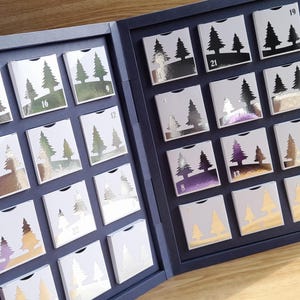 Könnte beinhalten: Geöffneter, marineblauer Adventskalender mit 24 kleinen, nummerierten Kästchen. Jedes Kästchen zeigt ein silbernes Baumdesign auf weißem Hintergrund. Die Kästchen sind in einem Raster innerhalb des Kalenderrahmens angeordnet.