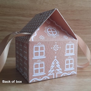 May include: A decorative gingerbread house-shaped gift box with a light brown color and white accents. The box features a roof, windows, and a Christmas tree design. A light brown ribbon is attached to the box.