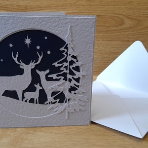May include: A gray card with a white, laser-cut design of a deer family and a pine tree in a circle against a dark blue background with white stars.