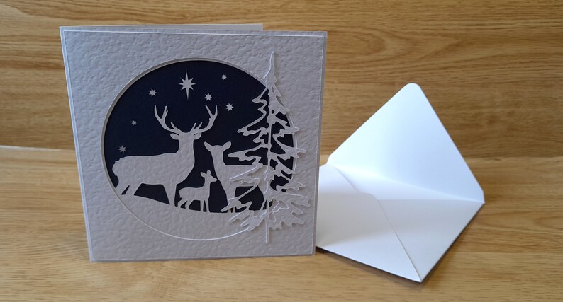 May include: A gray card with a white, laser-cut design of a deer family and a pine tree in a circle against a dark blue background with white stars.