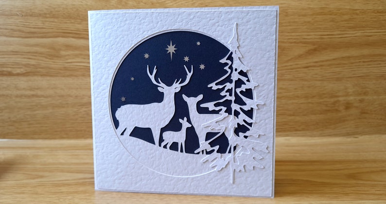 May include: A white card with a blue circle cut out in the center. Inside the circle are three white deer silhouettes and a pine tree silhouette. The background of the circle is dark blue with white stars.