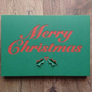 Op de afbeelding: Een groene kerstkaart met de woorden "Merry Christmas" uitgesneden in rood. Onder de tekst staat een uitgesneden ontwerp van hulstbladeren. De kaart ligt op een houten oppervlak.