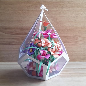 May include: A white geometric terrarium with a clear lid filled with a bouquet of pink, orange, and white paper flowers.