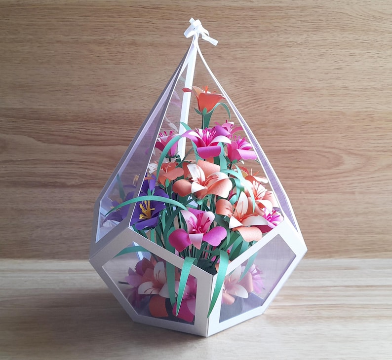 May include: A white geometric terrarium with a clear lid filled with a bouquet of pink, orange, and white paper flowers.