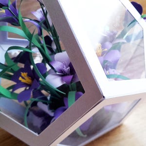 May include: A bouquet of purple and white paper flowers with green stems is displayed inside a clear, hexagonal box with a silver frame.