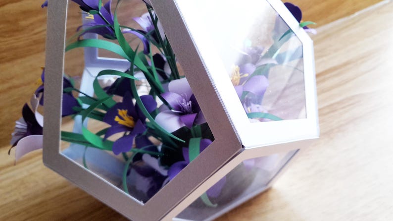 May include: A bouquet of purple and white paper flowers with green stems is displayed inside a clear, hexagonal box with a silver frame.