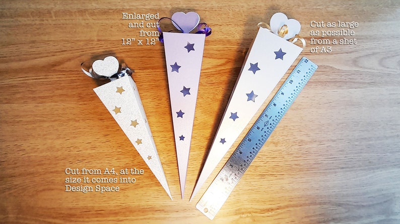 May include: Three cone-shaped paper party favor boxes with star cutouts. The boxes are made from white, light purple, and silver cardstock. The boxes are 12 inches by 12 inches, and the largest box is cut from a sheet of A3 paper. The smaller boxes are cut from A4 paper. The text "Cut from A4, at the size it comes into Design Space" is written below the silver box. The text "Enlarged and out from 12" x 12" is written above the light purple box. The text "Cut as large as possible from a sheet of A3" is written above the white box.