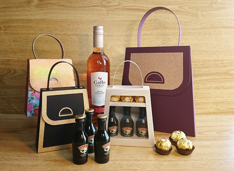 Pode incluir: Tr&ecirc;s sacolas de presente com al&ccedil;as, uma com uma garrafa de vinho branco Grenache da Gallo Family Vineyards, outra com uma caixa de chocolates e a &uacute;ltima com quatro garrafas pequenas de Baileys Irish Cream. As sacolas s&atilde;o decoradas com glitter dourado e t&ecirc;m um design de bolsa.