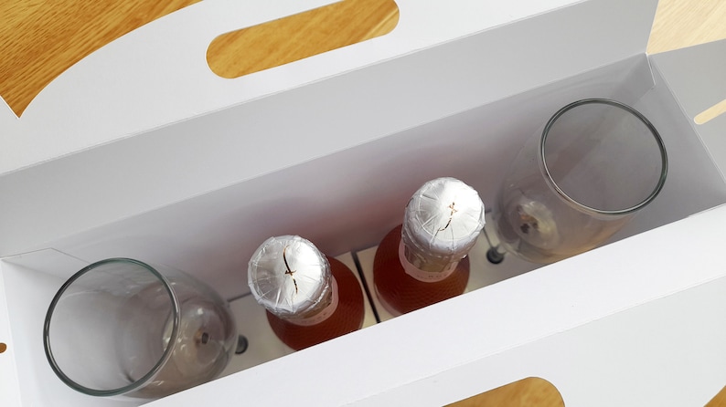 May include: A white gift box with a handle, containing two bottles of pink wine with silver foil tops and two clear glass wine glasses.