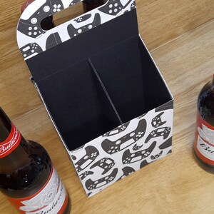 K&ouml;nnte beinhalten: Ein Sechs-Pack-Biertr&auml;ger aus Pappe mit wei&szlig;em Hintergrund und einem schwarz-wei&szlig;en Muster von Videospielcontrollern. Der Tr&auml;ger hat einen Griff und zwei F&auml;cher. Zwei Flaschen Budweiser-Bier stehen auf einer Holzoberfl&auml;che.