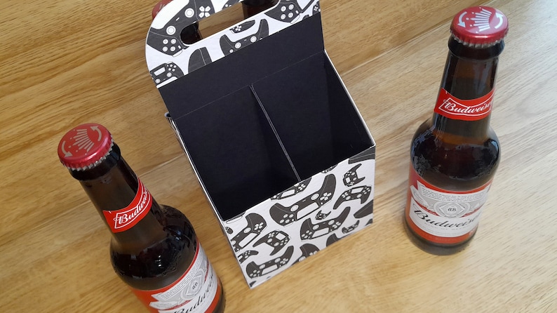 K&ouml;nnte beinhalten: Ein Sechs-Pack-Biertr&auml;ger aus Pappe mit wei&szlig;em Hintergrund und einem schwarz-wei&szlig;en Muster von Videospielcontrollern. Der Tr&auml;ger hat einen Griff und zwei F&auml;cher. Zwei Flaschen Budweiser-Bier stehen auf einer Holzoberfl&auml;che.