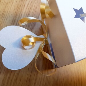 May include: A white gift box with a gold ribbon tied in a bow. The box has two star-shaped cutouts and a heart-shaped tag attached to the ribbon.