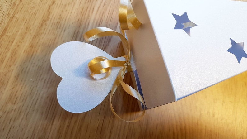 May include: A white gift box with a gold ribbon tied in a bow. The box has two star-shaped cutouts and a heart-shaped tag attached to the ribbon.