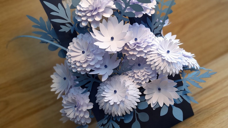 May include: A bouquet of paper flowers in shades of blue and white. The flowers are made of paper and have a 3D effect. The flowers are arranged in a vase and are surrounded by green leaves.