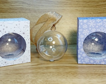 Op de afbeelding: Drie transparante glazen kerstballen in verschillende gepatroneerde dozen. De dozen zijn wit met een zwart sneeuwvlokkenpatroon, blauw met een wit sneeuwvlokkenpatroon en wit met een zwart bloemenpatroon.