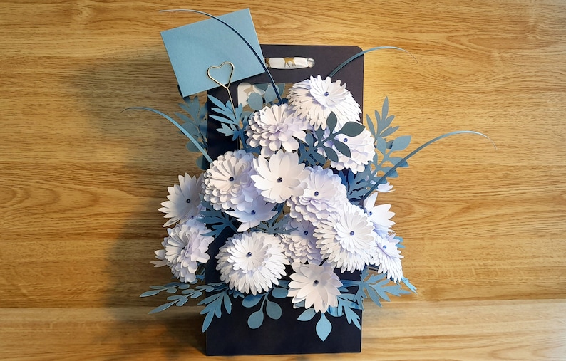May include: A paper craft bouquet of white flowers with blue paper leaves in a dark blue vase. A blue card with a gold heart paper clip is attached to the vase.