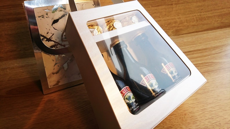 Pode incluir: Uma caixa de presente branca com uma janela transparente, contendo tr&ecirc;s garrafas em miniatura de Baileys Irish Cream e algumas trufas de chocolate. A caixa est&aacute; sobre uma superf&iacute;cie de madeira.