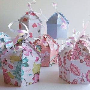 Puede incluir: Cinco pequeñas cajas de regalo decorativas con bordes festoneados. Las cajas están hechas de papel y tienen una variedad de patrones florales en rosa, azul y verde. Cada caja tiene una cinta atada alrededor.