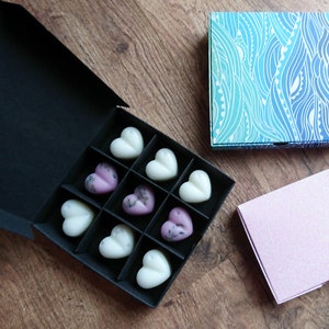 May include: A black box with nine heart-shaped wax melts, some white and some lavender. Two decorative boxes are also present, one with a blue and white wave pattern and another pink box with a glittery finish. The items are on a wooden surface.