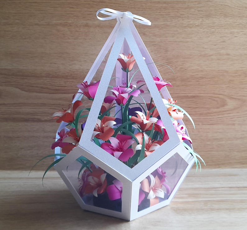 May include: A white geometric papercraft box with a clear lid, filled with pink, orange, and white paper flowers. The box has a white ribbon tied around the top.