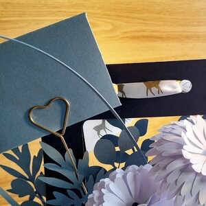 May include: A gray card with a gold heart-shaped paper clip, surrounded by paper flowers and leaves in shades of blue and purple. The card is blank and ready for a message.