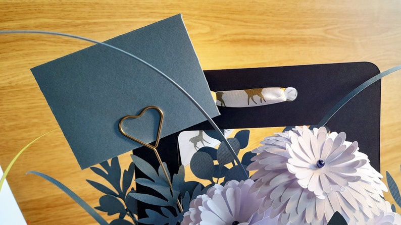 May include: A gray card with a gold heart-shaped paper clip, surrounded by paper flowers and leaves in shades of blue and purple. The card is blank and ready for a message.