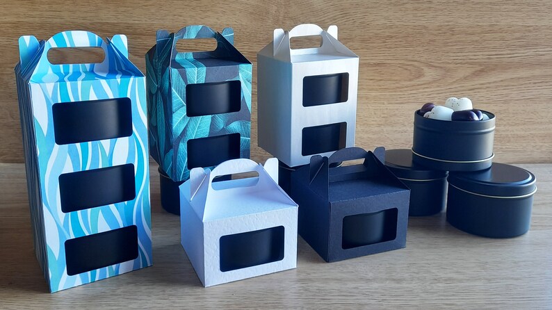 May include: Assortment of black candle tins with lids, packaged in various cardboard boxes with cut-out windows and handles. The boxes have different patterns, including blue waves, green leaves, and solid white and black.