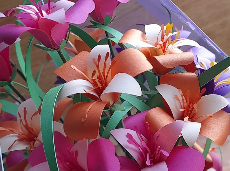 May include: A bouquet of paper flowers in shades of pink, orange, and white. The flowers are made of paper and have a realistic appearance. The bouquet is arranged in a clear vase.
