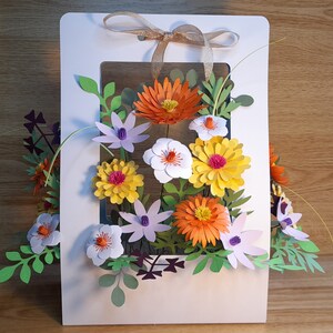 May include: A white card with a window cut out to display a paper flower arrangement. The flowers are made of paper and are in shades of orange, yellow, white, and purple. The flowers are arranged in a bouquet and are surrounded by green leaves.