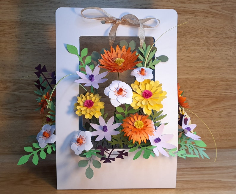 May include: A white card with a window cut out to display a paper flower arrangement. The flowers are made of paper and are in shades of orange, yellow, white, and purple. The flowers are arranged in a bouquet and are surrounded by green leaves.