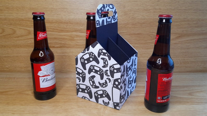 K&ouml;nnte beinhalten: Ein Sechs-Pack-Biertr&auml;ger aus Pappe mit wei&szlig;em Hintergrund und einem schwarz-wei&szlig;en Muster aus Videospiel-Controllern. Der Tr&auml;ger hat einen Griff oben und ist f&uuml;r sechs Bierflaschen ausgelegt. Drei Flaschen Budweiser-Bier sind auf dem Bild zu sehen.