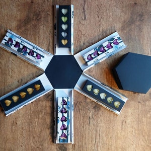 May include: An open, navy blue hexagonal gift box with six compartments, each holding heart-shaped chocolates in various colors. The box is decorated with floral patterns and sits on a wooden surface. A separate, closed navy blue hexagonal lid is also present.