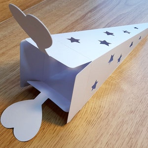 May include: White paper cone-shaped box with a heart-shaped flap on each end. The box has cut-out stars on the sides.