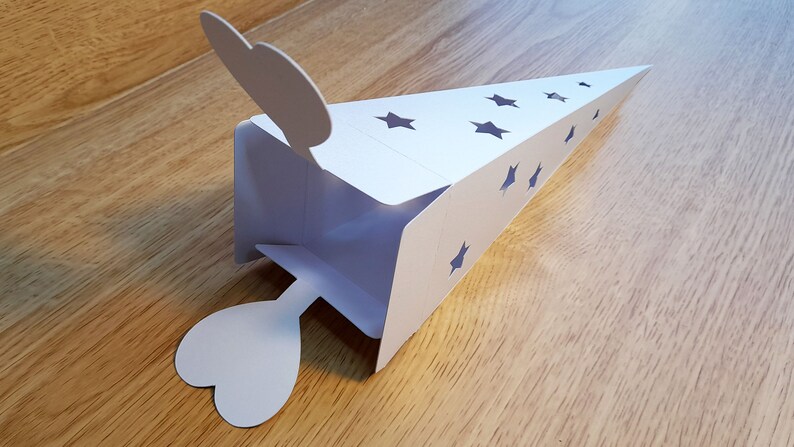 May include: White paper cone-shaped box with a heart-shaped flap on each end. The box has cut-out stars on the sides.