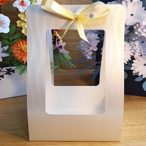 May include: A white paper gift box with a window and a yellow ribbon. The box is empty and has a white background.
