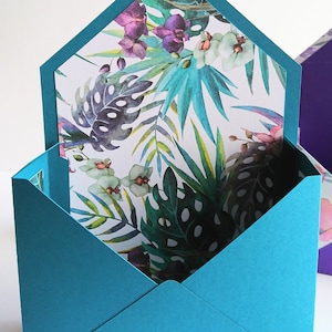 Könnte beinhalten: Ein türkisfarbener Umschlag mit einem weißen Futter, das ein tropisches Blumenmuster mit grünen Blättern, rosa und violetten Orchideen und weißen Blüten zeigt.