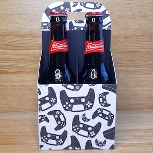 May include: A six-pack carrier with a white background and a black and white pattern of video game controllers. The carrier has two brown glass bottles of Budweiser beer inside.