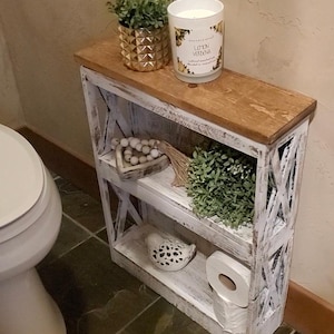 May include: A narrow, two-shelf wooden storage unit with a distressed white finish and a natural wood top. The unit holds a gold geometric planter with greenery, a Lemon Verbena candle, wooden beads, a white bird figurine, and toilet paper.