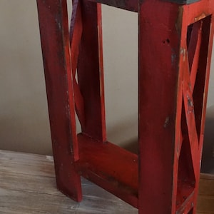 May include: A tall, narrow wooden stand featuring a distressed red painted finish on its frame and a dark, weathered rectangular top. The structure includes vertical supports connected by cross-bracing on the sides and a lower horizontal shelf.