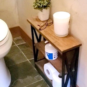 May include: A narrow, two-tier wooden shelving unit with a dark metal X-frame design. The top shelf holds a small potted green plant, a pair of brown-rimmed glasses, and a white candle. The bottom shelf contains a white container and toilet paper rolls.