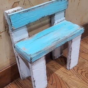 May include: A small bench constructed from reclaimed wood, featuring a distressed finish with bright turquoise paint on the seat and backrest, contrasted by white painted legs and support structures, showcasing a weathered and rustic wood grain texture.