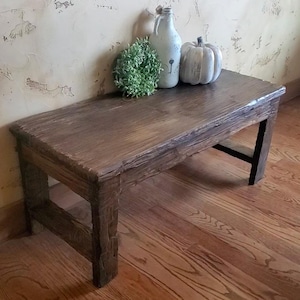 May include: A rectangular wooden bench with a dark brown stained finish and visible wood grain texture. The bench features four sturdy legs and a flat top surface. Resting on the bench are a green faux plant, a white bottle, and a white pumpkin.