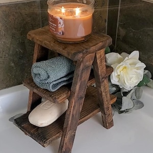 May include: A rustic, three-tiered wooden shelf unit. A lit candle in a glass jar sits on top, with a folded blue towel and a bar of soap on the lower shelves. White flowers and greenery are to the side.